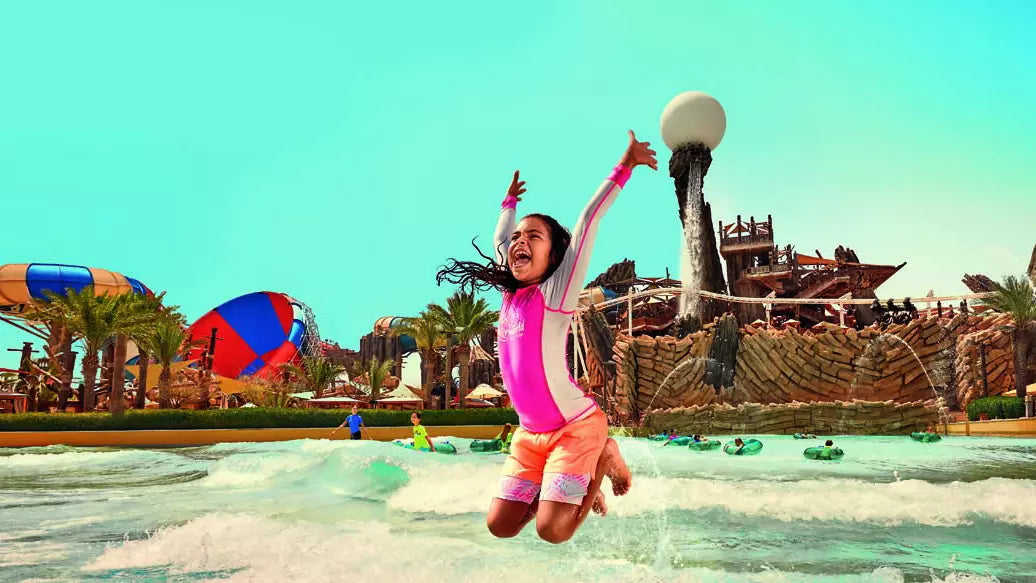 Child jumping in a water park with a colorful slide and tropical resort in the background